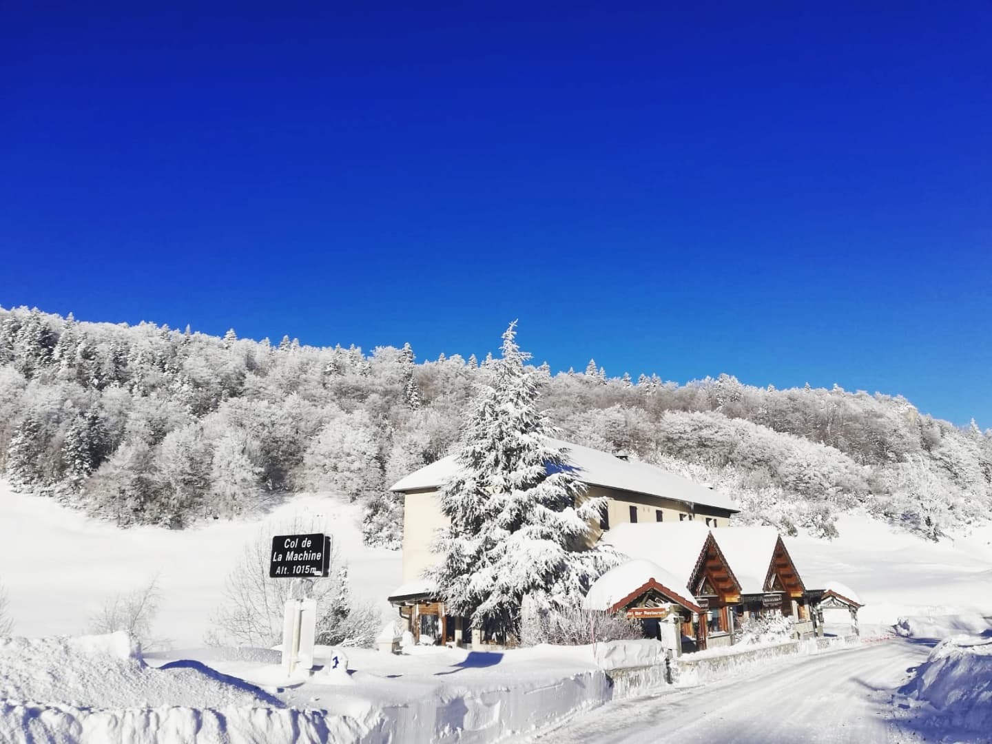 Le panneau du col de la Machine (alt. 1015 m) se dresse dans un décor enneigé où route, établissement et sapins se trouvent sous un ciel lumineux