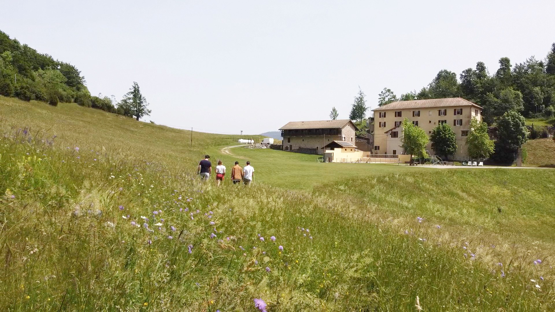 Au premier plan, une prairie fleurie que traversent quatre personnes vues de dos ; en arrière-plan, à droite, l’auberge apparaît, visible depuis l’arrière
