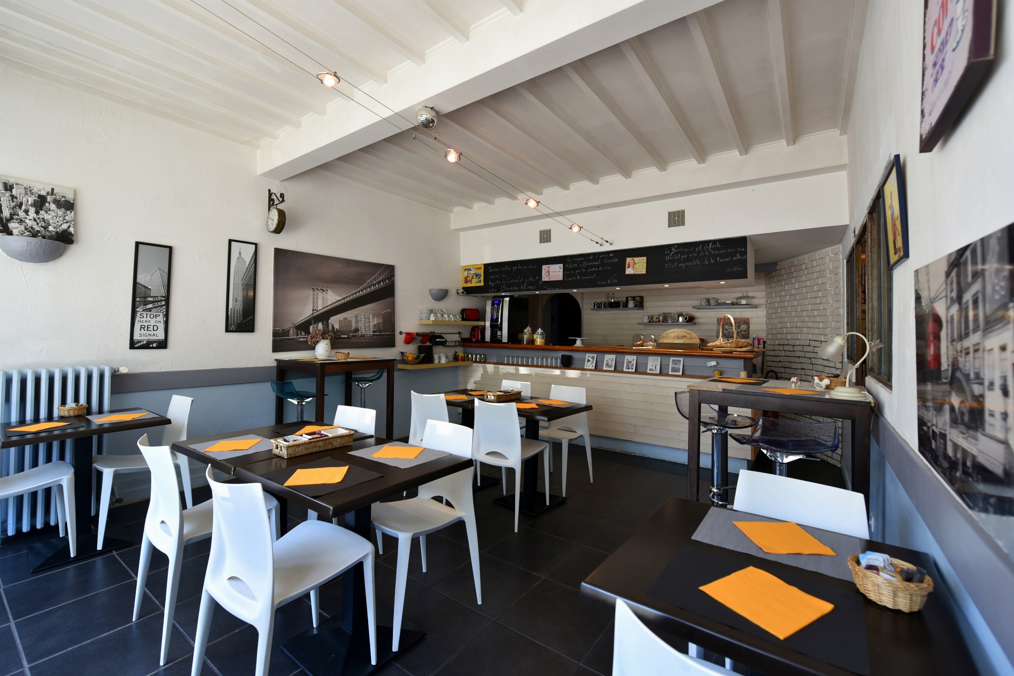 Salle de petit déjeuner avec six tables dressées dont deux tables hautes pour deux personnes. Un buffet petit déjeuner est installé en arrière plan. Les murs sont décorés de nombreux cadres et d’une horloge