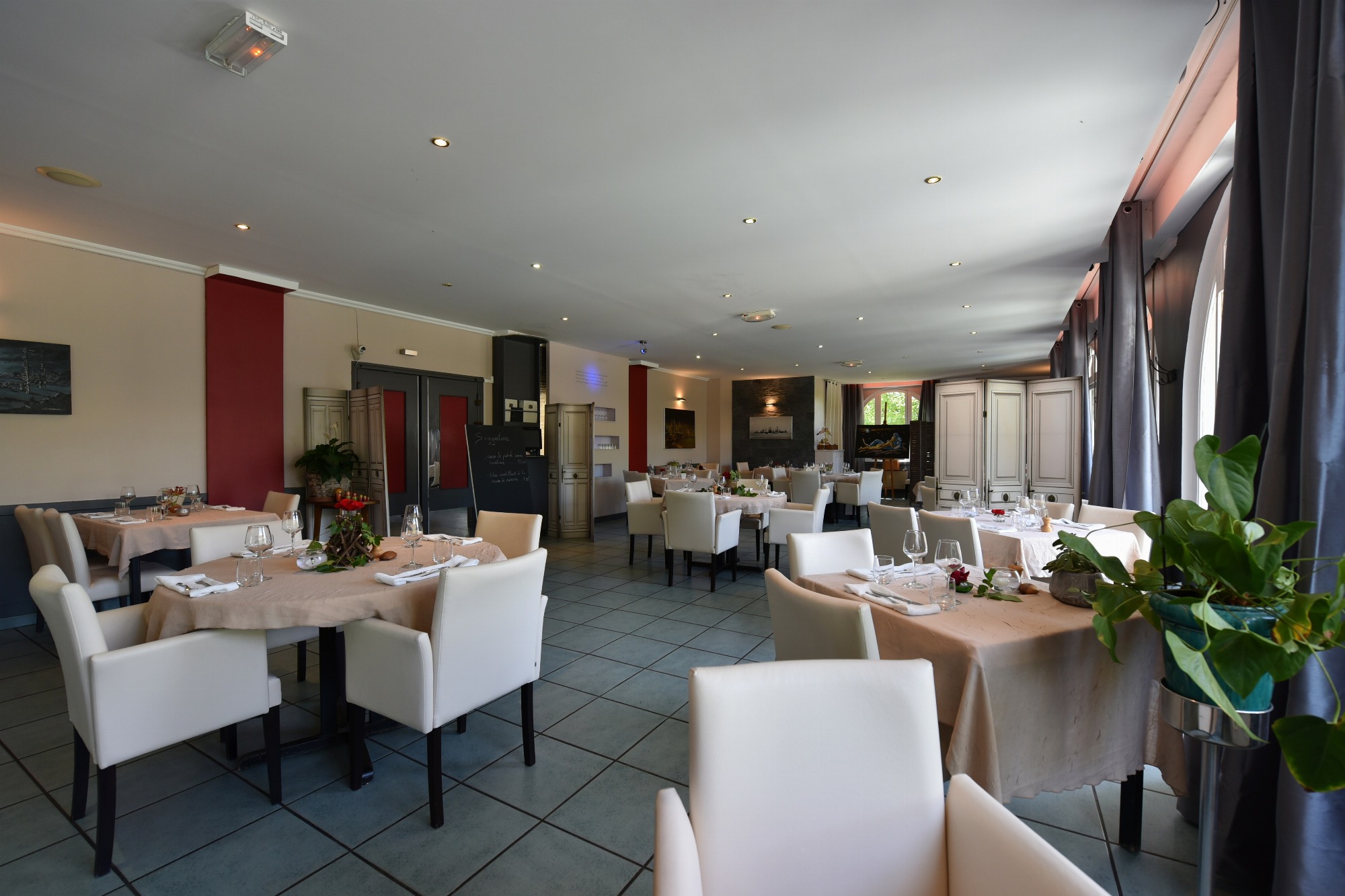 Une dizaine de tables sont dressées pour le repas dans une salle de restauration spacieuse décorée avec des tableaux et des plantes vertes