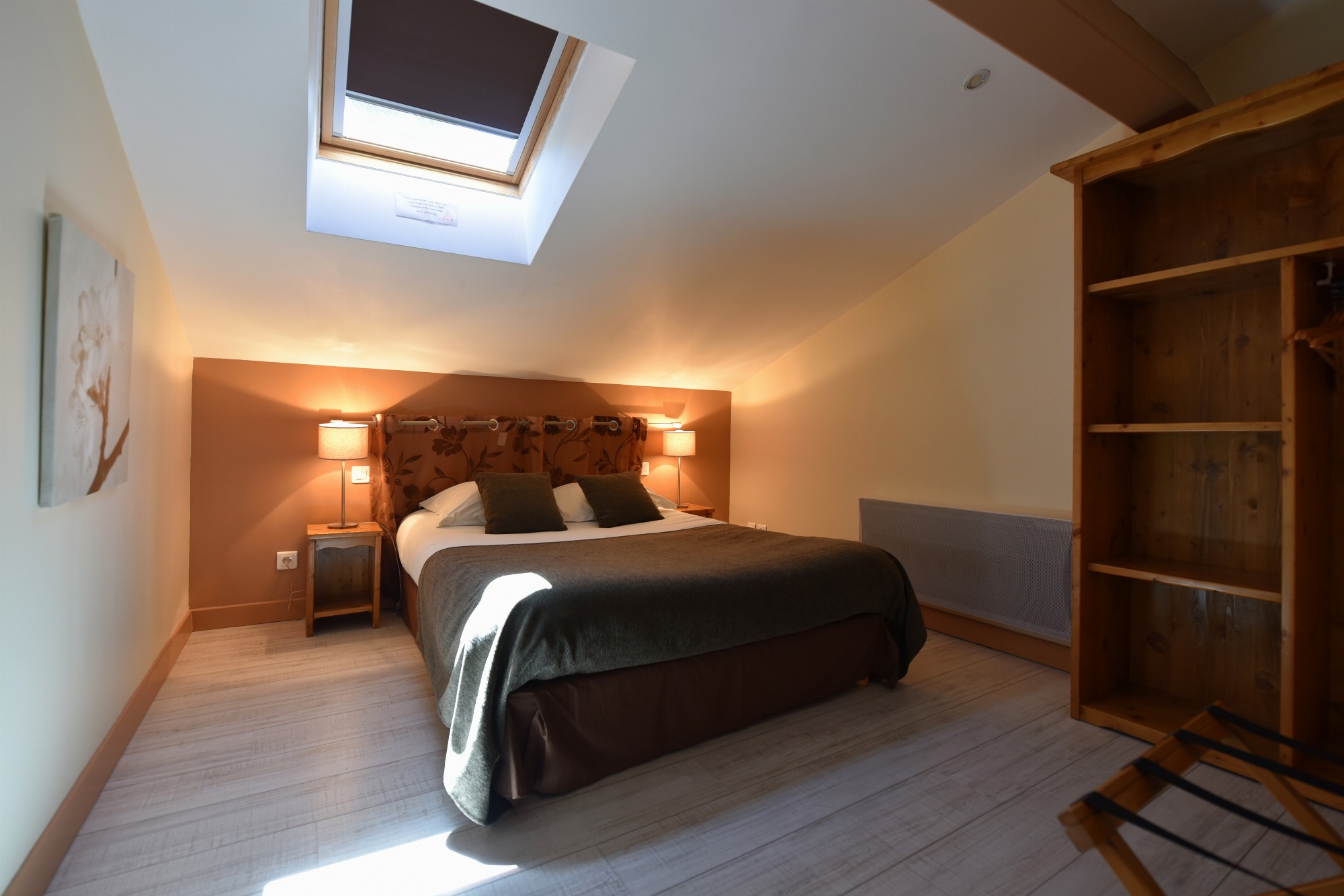 Chambre dotée d’un lit double, de deux tables de chevet avec lampes allumées, d’un vélux laissant entrer la lumière naturelle, d’un cadre mural, d’une armoire ouverte et d’un porte-bagage. Un radiateur est aussi visible dans la pièce