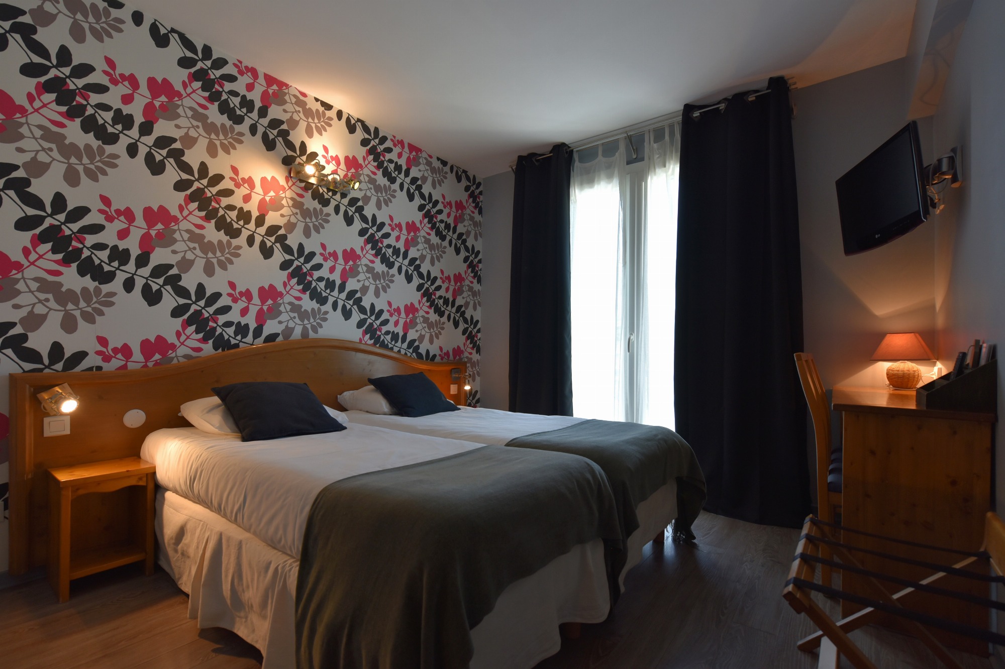 Chambre avec deux lits simples, chacun accompagné d’une table de chevet éclairée par un spot. Une applique murale et une lampe de bureau complètent l’éclairage. Présence d'un téléviseur et d'un porte bagage