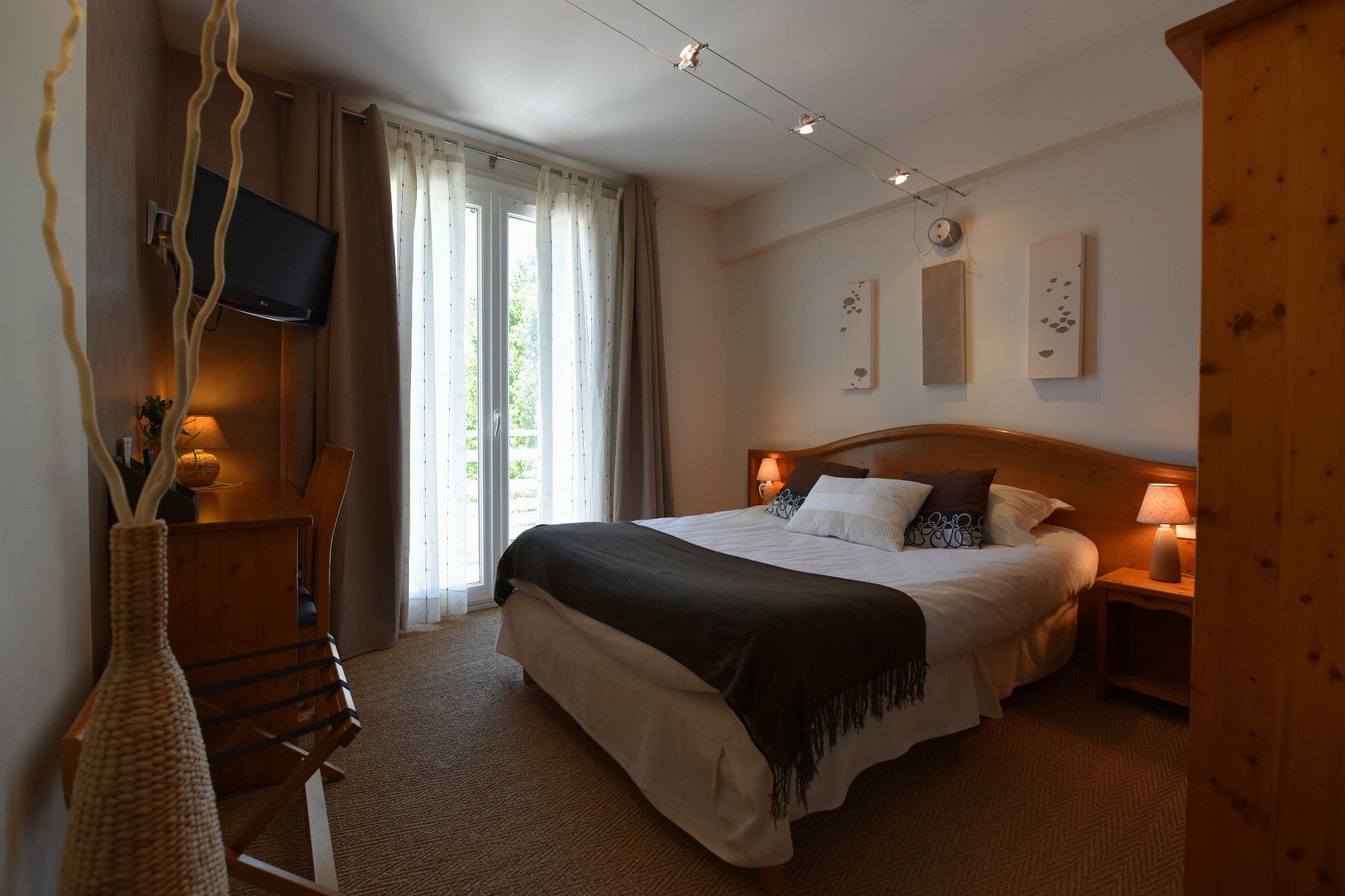 Chambre double chaleureuse équipée de deux lampes de chevet et d’une lampe de bureau allumées, de trois cadres décoratifs au-dessus du lit, d’une armoire, d’un porte bagage et d’un téléviseur. Trois spots orientables éclairés sur rail assurent un éclairage central. Vue sur la verdure par la porte-fenêtre