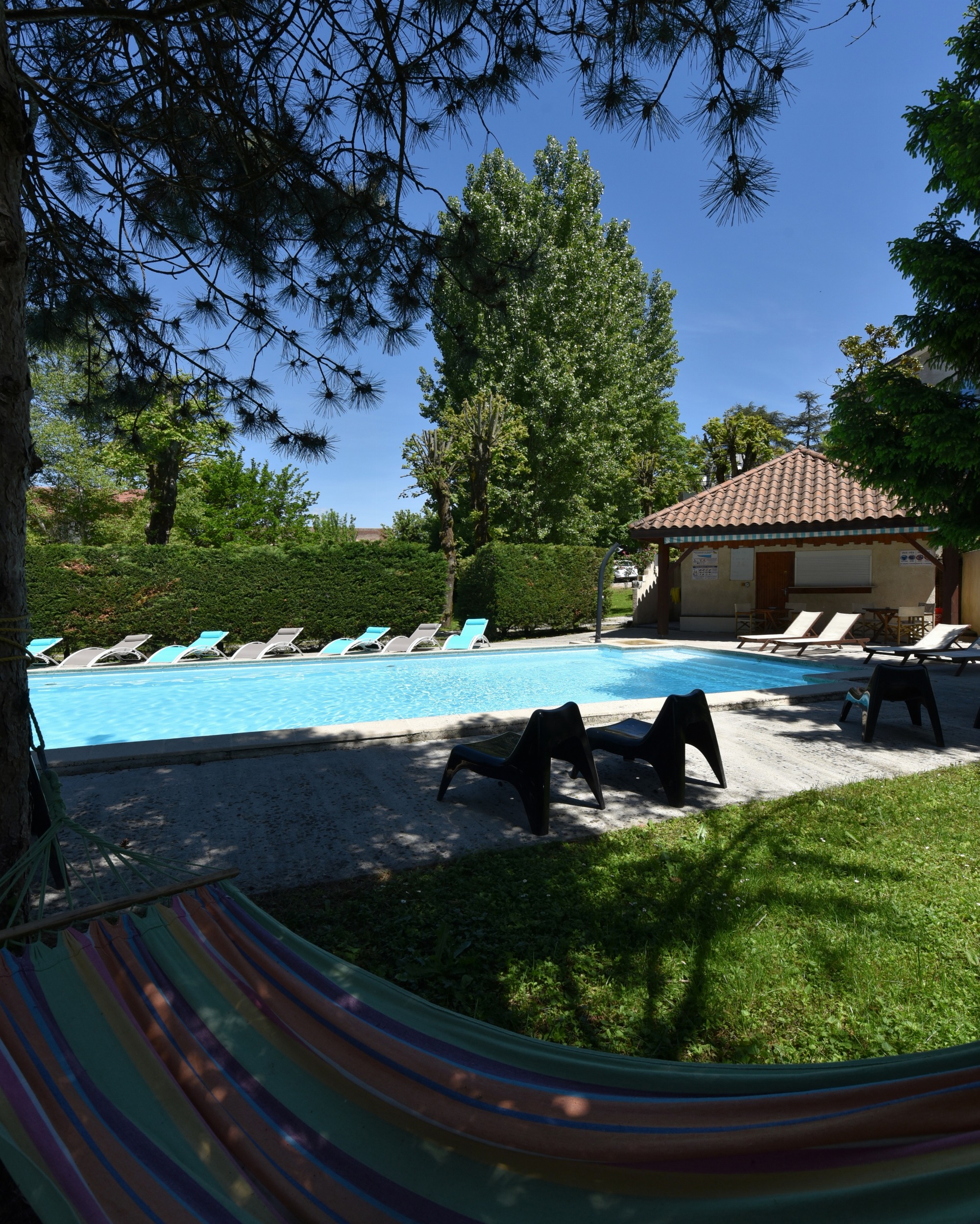Espace piscine entouré d’arbres et comprenant des transats, des sièges, un hamac en premier plan, le tout installé devant le local technique