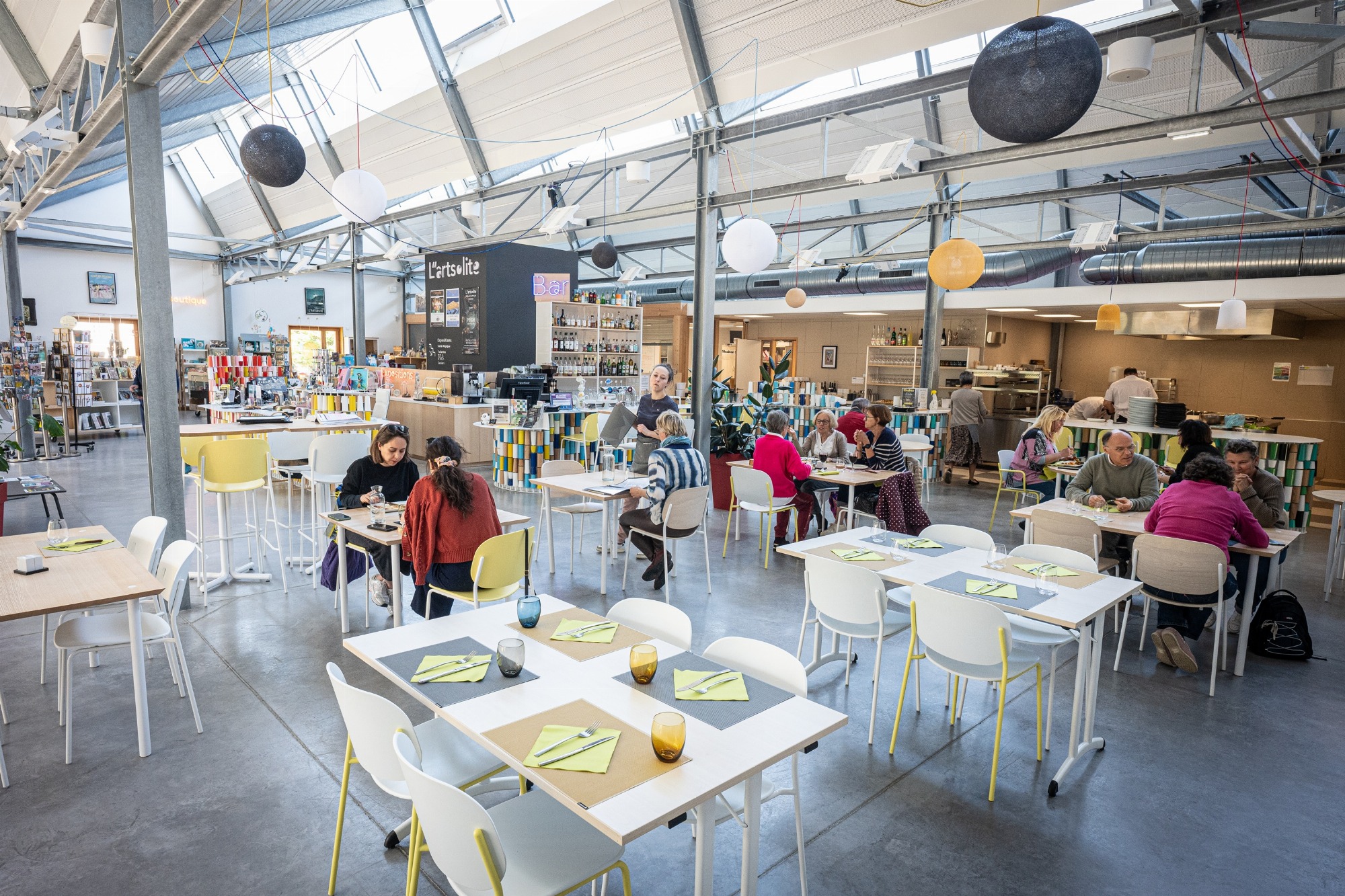 Des convives sont installés dans la salle de restauration avec en arrière plan les espaces cuisine, bar et boutique